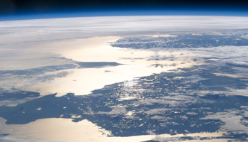 View of Earth from space showing a sunlit coastline, clouds, and the blue curvature of the planet's atmosphere.