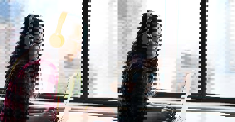 A woman with long hair wearing a red check shirt and orange headphones sitting at a laptop in front of a window